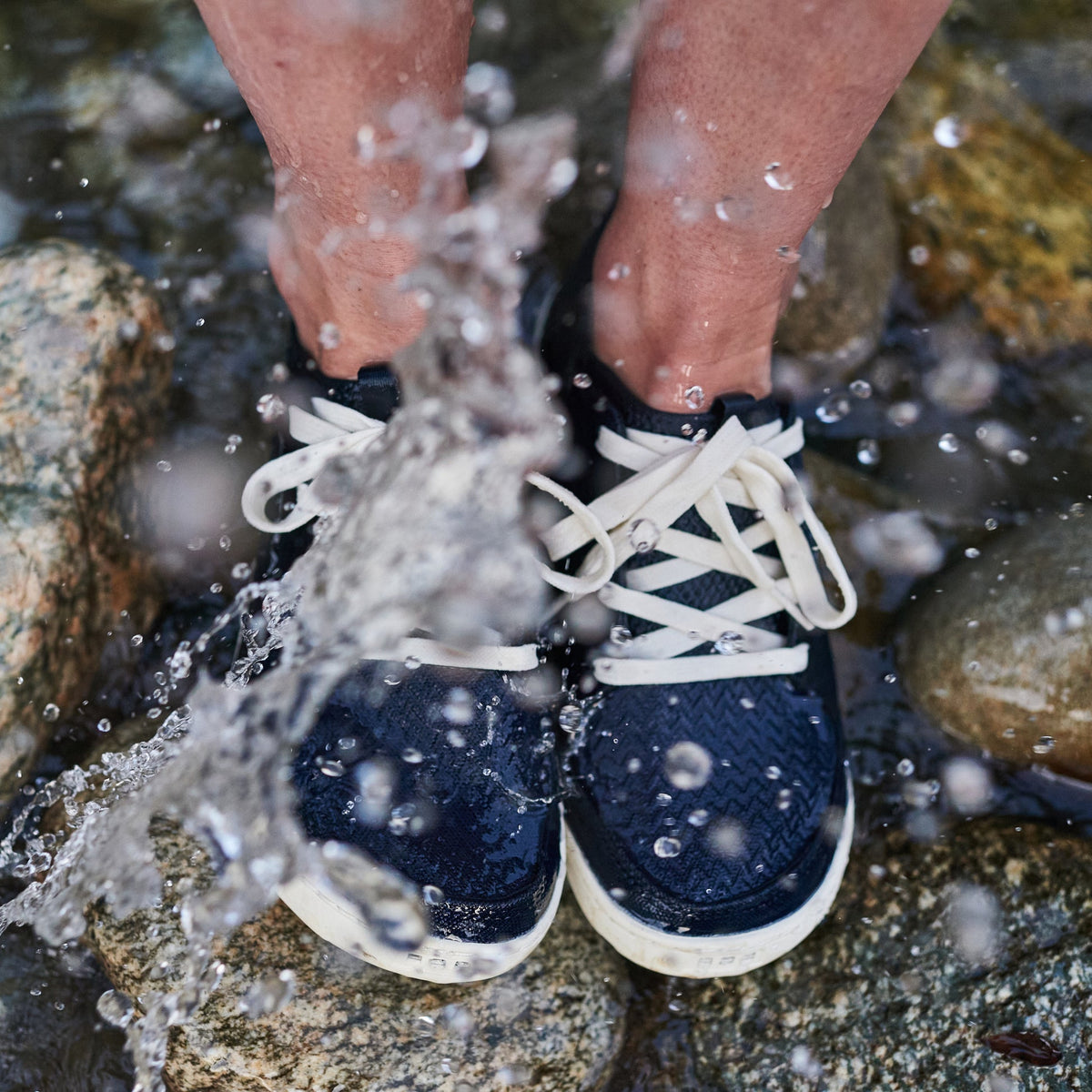 Close-up of Astral Loyak W water shoes in navy with white laces, feet immersed in a rocky stream with splashing water.