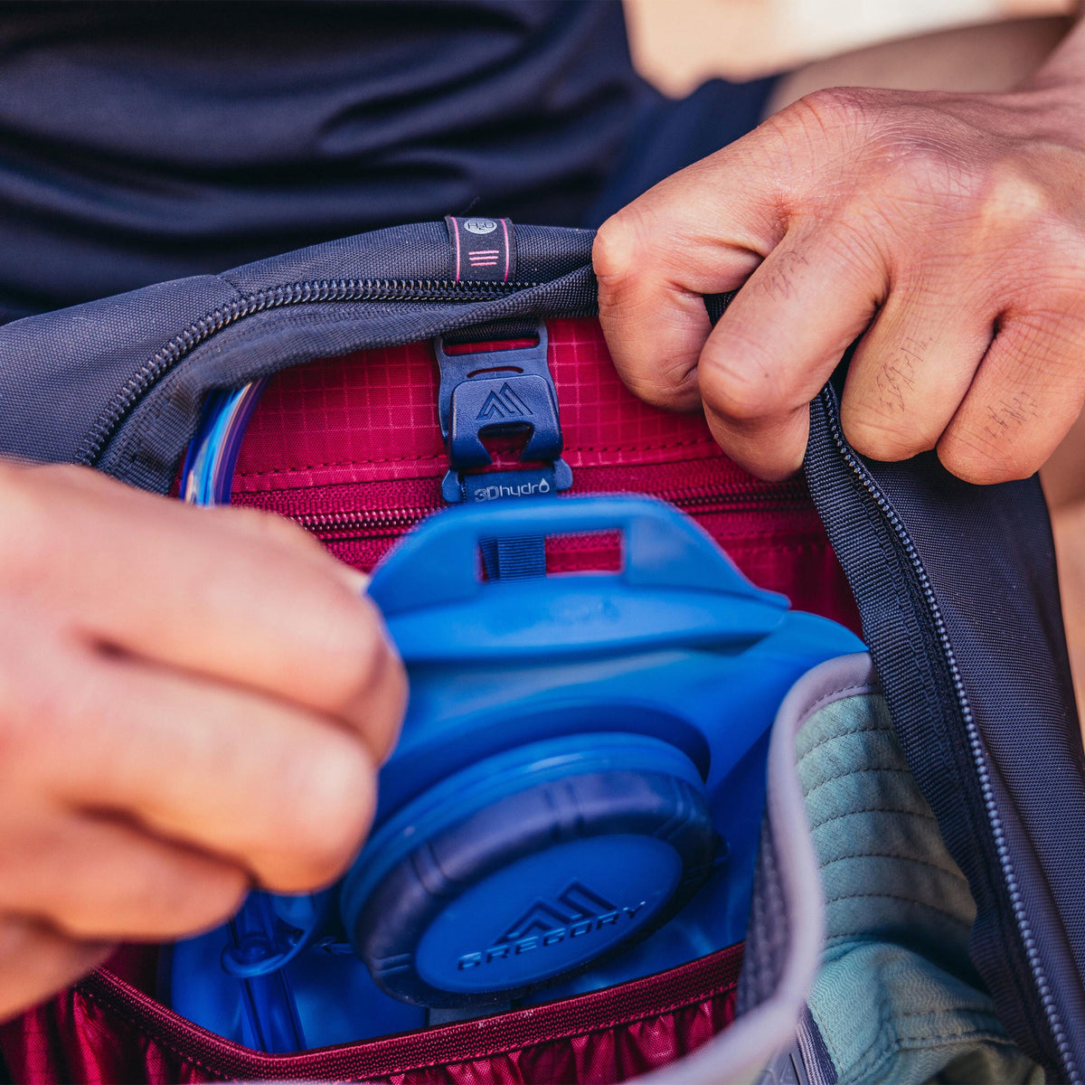 Hands placing a blue Gregory 3Dhydro hydration bladder into the red internal compartment of a Gregory Zulu 24 LT backpack.