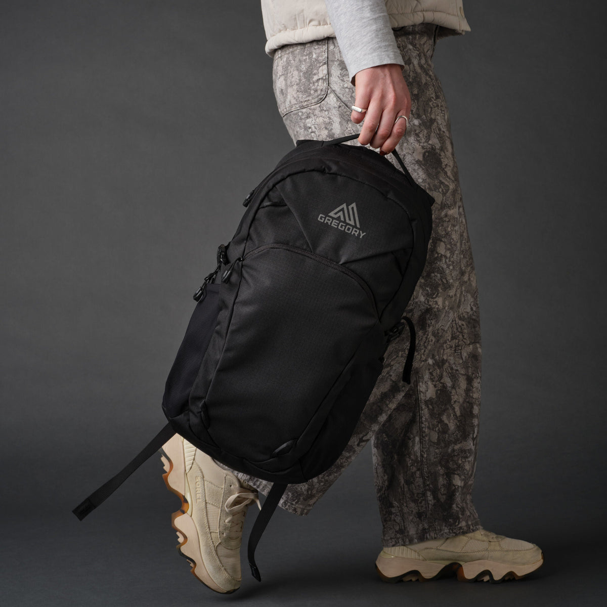 Side view of a person walking, carrying a black Gregory Signal 18 daypack in hand, wearing patterned pants and beige sneakers.