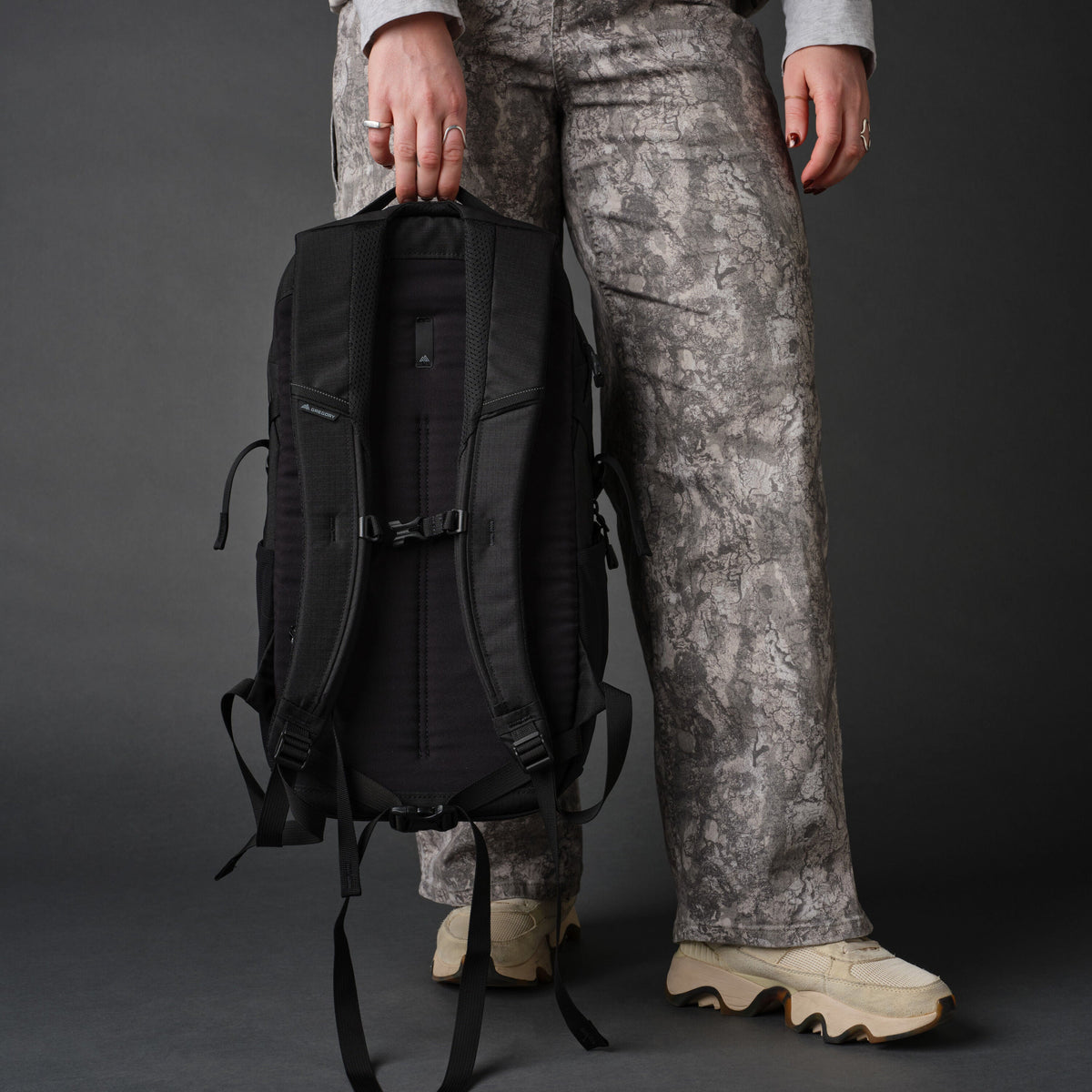 Person holding a black Gregory Signal 18 backpack by the top handle, wearing grey patterned pants and cream sneakers.