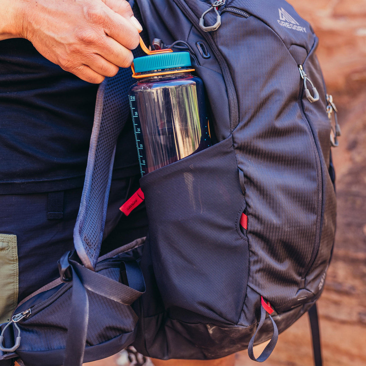 Hiker's hand reaching for a Nalgene water bottle in the mesh side pocket of a dark grey Gregory Zulu 24 LT backpack.