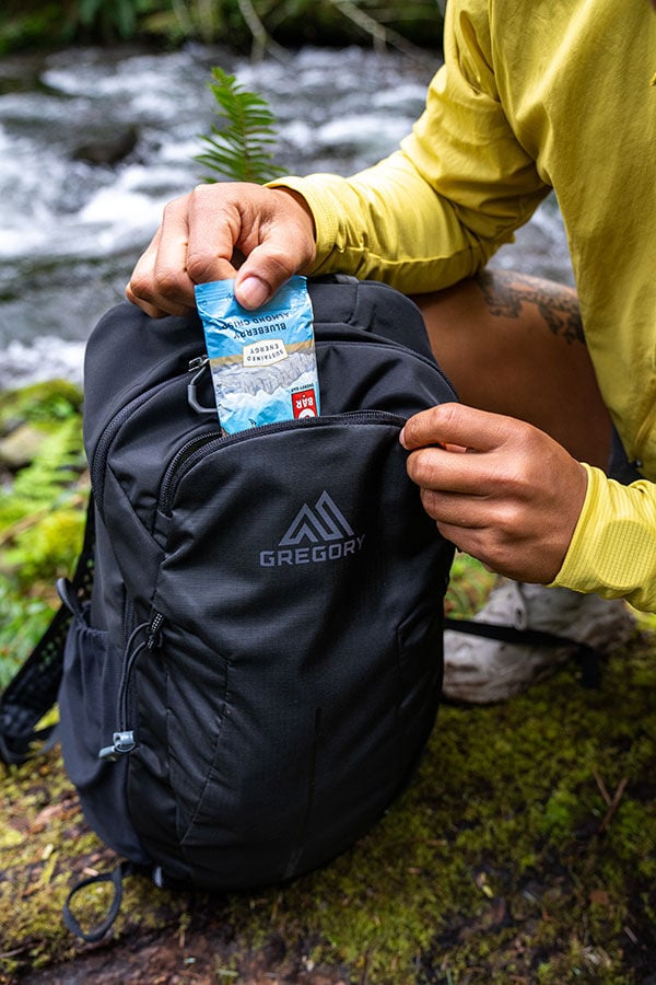 Person putting a snack bar into a black Gregory Arrio 20 backpack in a natural outdoor setting by a river.